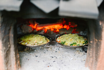 Two savory dishes with melted topping baking inside a rustic wood-fired oven with glowing embers, traditional cooking.