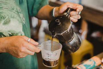 A close-up shot of an adult pouring hot, dark coffee from a classic moka pot into a clear measuring cup, emphasizing the brewing process and homemade ritual.