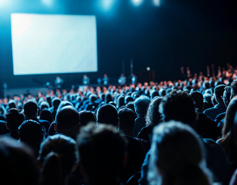 A large group of business people seated in an auditorium, attending a work conference presentation on stage with a big blank screen under blue lighting.