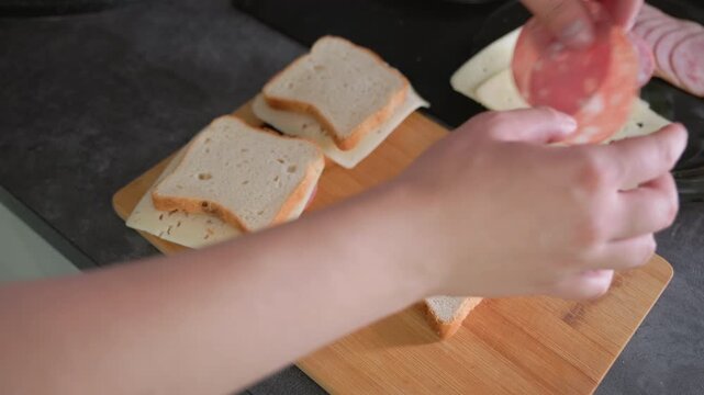 Hands assembling sandwiches with sliced cheese and cold cuts on wooden cutting board in kitchen. Several sandwiches partially made, ingredients like bread and meat placed neatly beside, in process of
