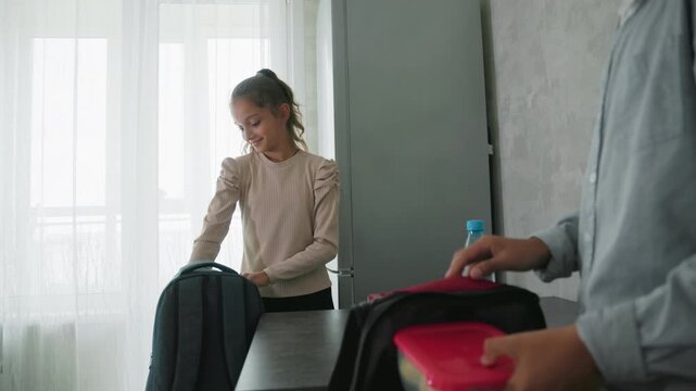 Girl and boy wearing casual clothes preparing for school by placing lunch containers and water bottles into backpacks beside kitchen table in brightly lit room with curtain-covered window and - Powered by Adobe