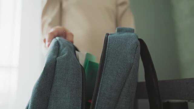 girl in casual beige sweater packing green notebook into blue school bag near window, preparing for class in soft morning light, organized school supplies visible, focused hand gesture emphasizes