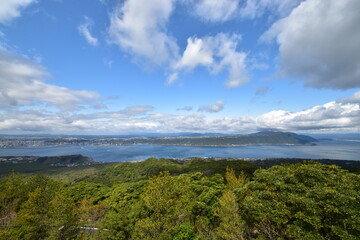 A breathtaking panoramic vista from Sakurajima island, Japan, featuring lush green forests in the foreground, the expansive Kinko Bay, and the city of Kagoshima under a blue sky.


