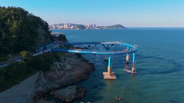 Aerial circular skywalk extending over the ocean from a rocky coastline. People are on the walkway with a distant city skyline, Busan, South Korea.