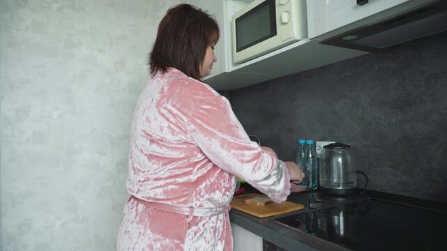 Woman in pink velvet robe stands in modern kitchen preparing sandwiches with sliced bread, cheese, meat, apples, and bottled water on counter, creating peaceful domestic moment in natural light - Powered by Adobe