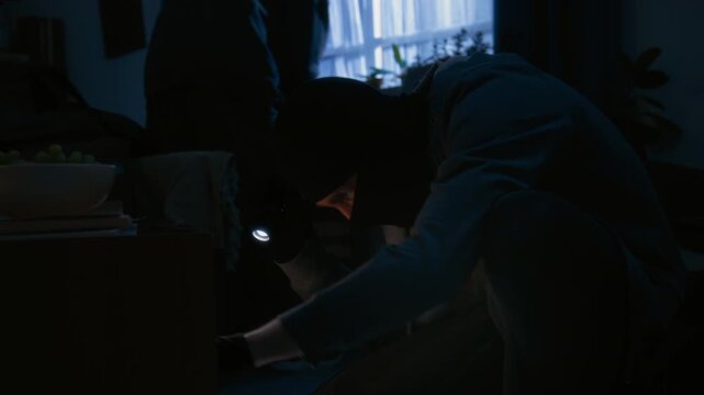 Unrecognizable thief in hoodie and black balaclava squatting near coffee table with shelf underneath and checking it with flashlight. He taking tablet and putting it in bag