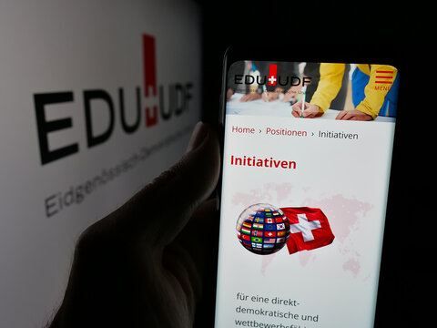 Stuttgart, Germany - 07-30-2025: Person holding cellphone with webpage of party Eidgenossisch-Demokratische Union (EDU) on screen in front of logo. Focus on center of phone display.