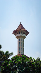 Fototapeta premium Tall mosque tower with traditional red tiled roof, surrounded by lush green trees under a clear blue sky.