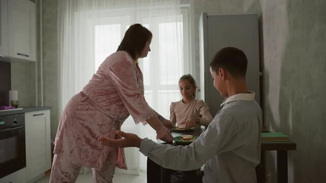 woman in pink robe serving sandwiches on plates to children at kitchen table, one child eagerly reaching for sandwich while other looks bored, bright morning light in modern home kitchen