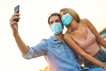 Young couple wearing protective face mask taking selfie at sunset