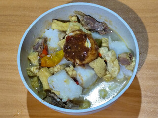 A bowl of vegetable lontong with coconut milk sauce and accompaniments of tofu, beef and Indonesian petis eggs, served on a wooden table.
