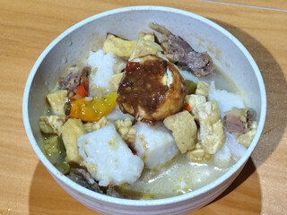 A bowl of vegetable lontong with coconut milk sauce and accompaniments of tofu, beef and Indonesian petis eggs, served on a wooden table.