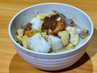A bowl of vegetable lontong with coconut milk sauce and accompaniments of tofu, beef and Indonesian petis eggs, served on a wooden table.