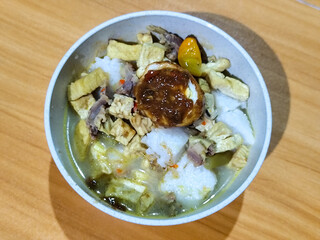 A bowl of vegetable lontong with coconut milk sauce and accompaniments of tofu, beef and Indonesian petis eggs, served on a wooden table.