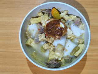 A bowl of vegetable lontong with coconut milk sauce and accompaniments of tofu, beef and Indonesian petis eggs, served on a wooden table.