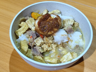 A bowl of vegetable lontong with coconut milk sauce and accompaniments of tofu, beef and Indonesian petis eggs, served on a wooden table.