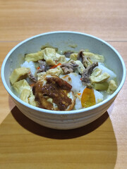 A bowl of vegetable lontong with coconut milk sauce and accompaniments of tofu, beef and Indonesian petis eggs, served on a wooden table.