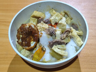 A bowl of vegetable lontong with coconut milk sauce and accompaniments of tofu, beef and Indonesian petis eggs, served on a wooden table.