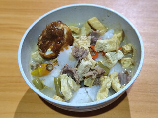 A bowl of vegetable lontong with coconut milk sauce and accompaniments of tofu, beef and Indonesian petis eggs, served on a wooden table.