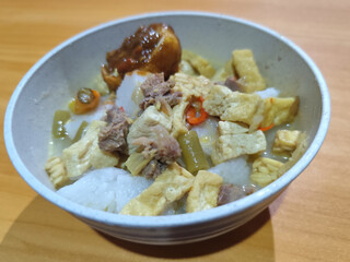 A bowl of vegetable lontong with coconut milk sauce and accompaniments of tofu, beef and Indonesian petis eggs, served on a wooden table.