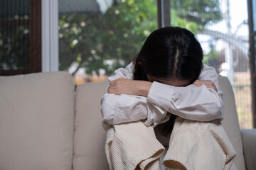 A person sitting on a couch, with their arms wrapped around their knees, appearing distressed or contemplative in a softly lit room.