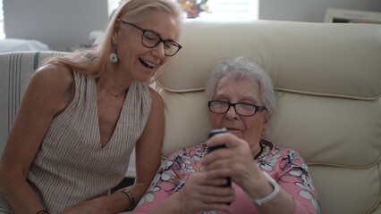 A smiling daughter supports her elderly mother in learning how to make a phone call while visiting a senior living facility, fostering connection and memory.