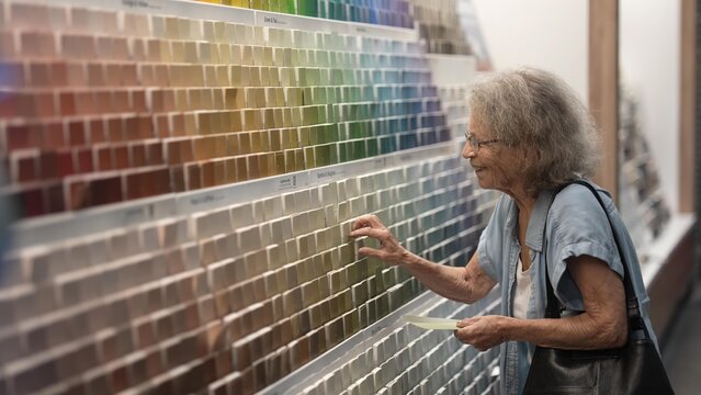 An elderly woman browses paint color samples in a hardware store, carefully considering her options for a home decorating project. She seems focused and thoughtful.
