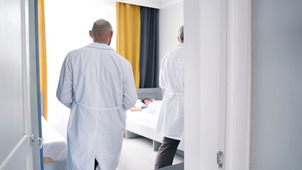 Fototapeta premium Two doctors enter the patient's room, view from the back. Two men in white coats are standing in front of a sick man's bed. Morning rounds of patients at the clinic