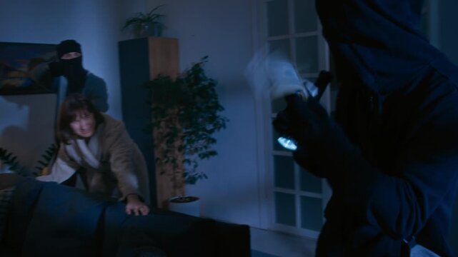 Burglar in black balaclava with flashlight in hands listening to scared woman who crying and pointing at wardrobe with money inside