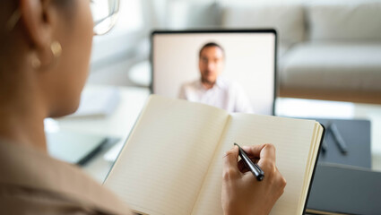 Close-up of a person writing in a notebook while attending an online video meeting with a blurred man on the laptop screen