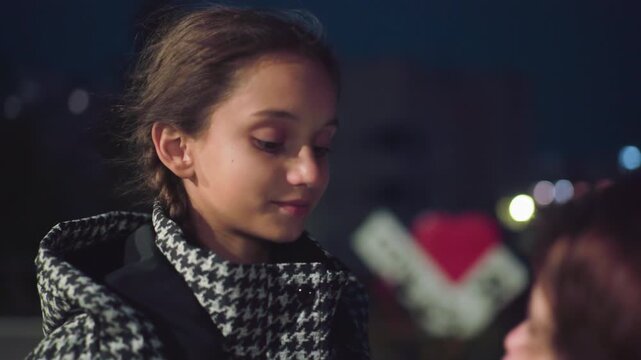 Young girl in houndstooth coat with subtle smile looks down thoughtfully at woman in soft evening light near urban background, capturing tender emotional moment filled with warmth, care, and quiet