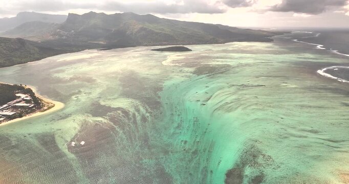 Aerial view of an underwater fall in the turquoise ocean with a beautiful reef and scenic mountains, Riviere Noire, Mauritius.