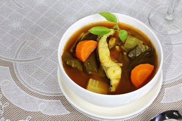 Thick sour curry with pieces of fish and vegetables in a bowl, garnished with fresh green leaves, served on a table with a white tablecloth.

