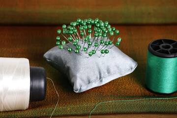 There are many pins stuck on a small grey pillow, which is placed between the white and green threads on a large piece of cloth.

