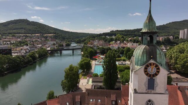 clocktower rises over rooftops as river curves toward olten cityscape under summer sky