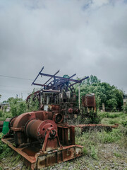 A wide shot of a huge, heavily rusted industrial machine, covered in vines and weeds, with a cab and large winches on a cloudy day