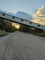 Obraz premium An industrial conveyor belt stretches across a sand-covered road with tire tracks, leading to a vibrant sunset peeking through green trees