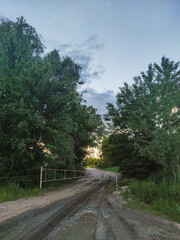 A muddy, rutted dirt road is blocked by a white and red striped barrier, leading into a dense line of trees with a bright, hazy sunset glow visible through the foliage under a dusky sky.