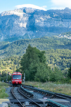A red train departing Servoz station in the French Alps with green hills and rocky mountains behind.