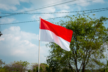 ndonesian red and white flag waving proudly against a blue sky with trees in the background, symbolizing patriotism and the celebration of Independence Day, August 17th.