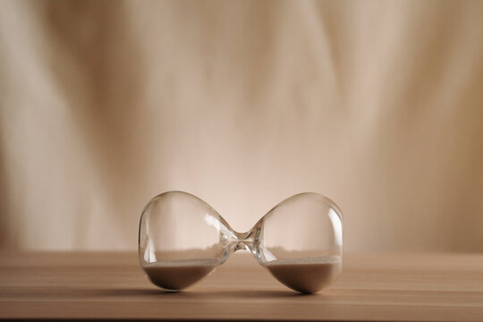 A simple hourglass on it's side on a wooden table with a beige backdrop, symbolizing time flow and minimalistic living. Represents timeless balance in a creative, uncluttered presentation concepts - Powered by Adobe