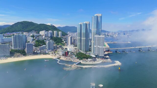 Aerial view of a modern coastal city, featuring skyscrapers, a beach, a bridge, and mountains, with fog rolling in from the sea in Busan, South Korea.