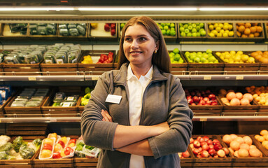 Confident female employee with arms crossed standing against shelves with fruits and vegetables in a grocery store