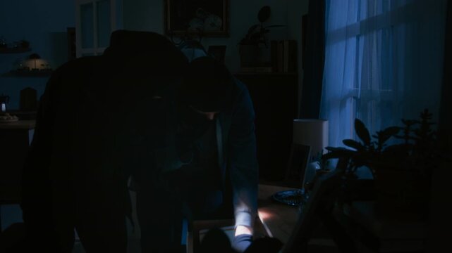 Two unrecognizable thieves in black balaclavas rummaging through drawer in living room during break in while police arriving