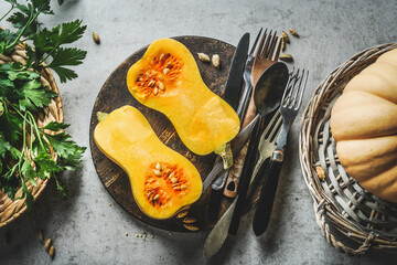 Halved butternut squash on plate with cutlery and herbs at grey concrete kitchen table background. Cooking at home with seasonal vegetables. Top view.