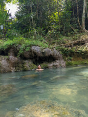 A natural landscape featuring a river flowing between large rocks. The water is clear and bluish, with small ripples. Many trees can be seen surrounding the river.