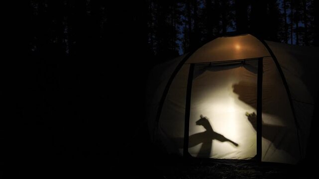 Silhouettes of children create playful shadow puppets inside a glowing tent during nighttime in the forest, capturing a fun and creative moment of outdoor adventure
