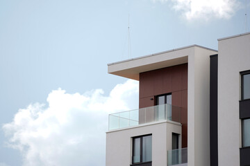 Modern apartment building corner with white and dark gray walls, featuring glass balconies and large windows. The upper part is accented with a wooden panel, set against a bright sky with fluffy cloud