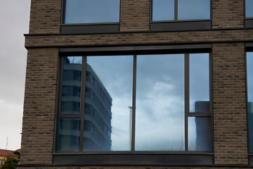 Fototapeta premium Reflection of a modern curved building and cloudy sky on the glass windows of a brick facade building. The windows show slight smudges, adding texture to the urban architectural scene.