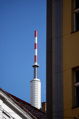 Obraz premium Tall communication tower with red and white stripes rises above rooftops against a clear blue sky. Architectural details of the building roof and chimney are visible. Clear daytime urban scene.